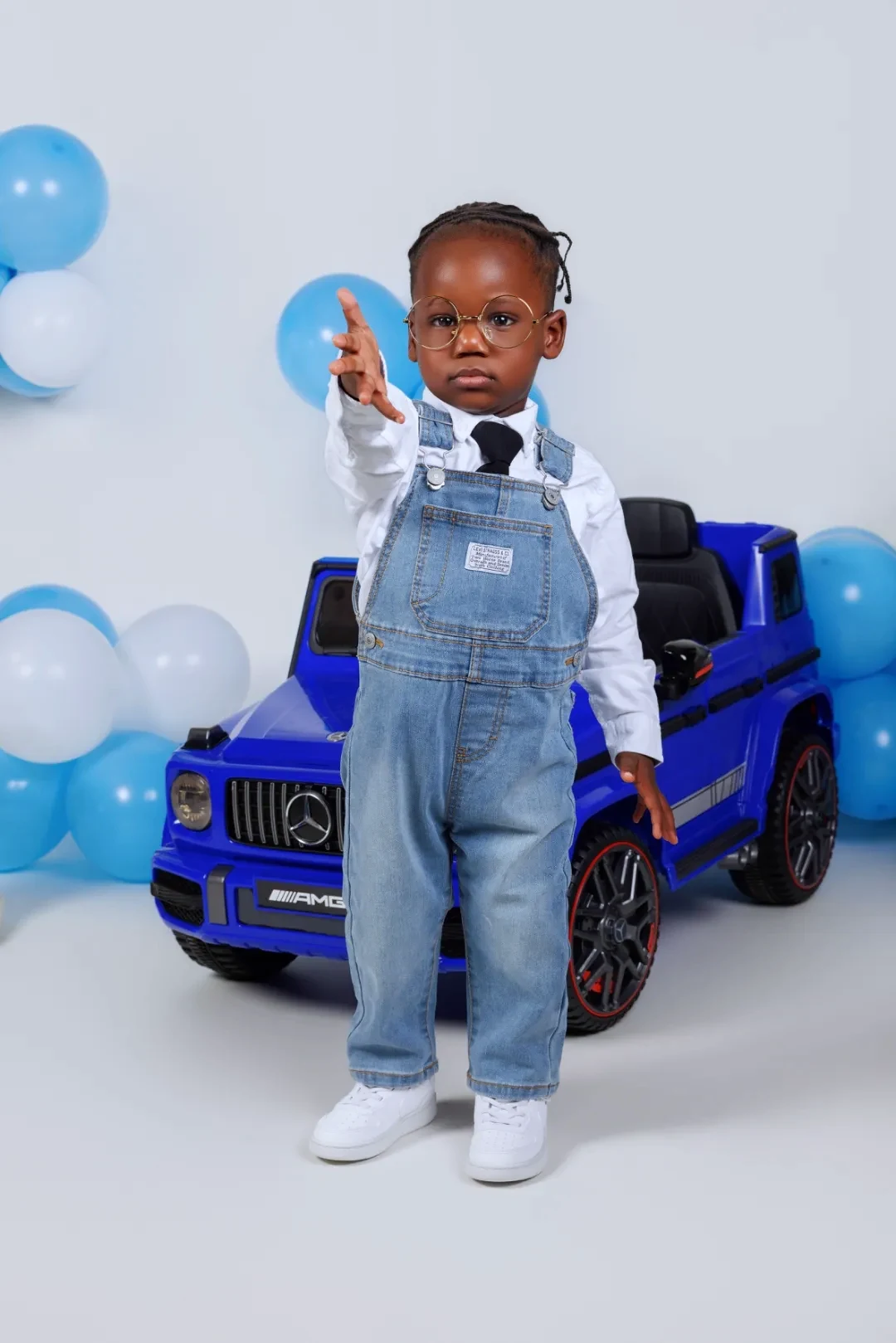 Toddler in denim overalls posing beside a toy ride-on car for a 2nd birthday shoot