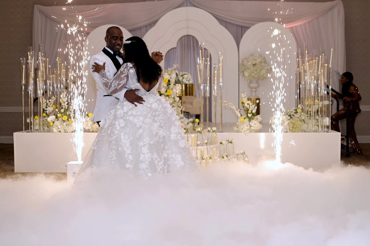 Bride and groom sharing their first dance surrounded by cold-spark fountains and low-lying dance floor fog, in front of a white floral arch backdrop