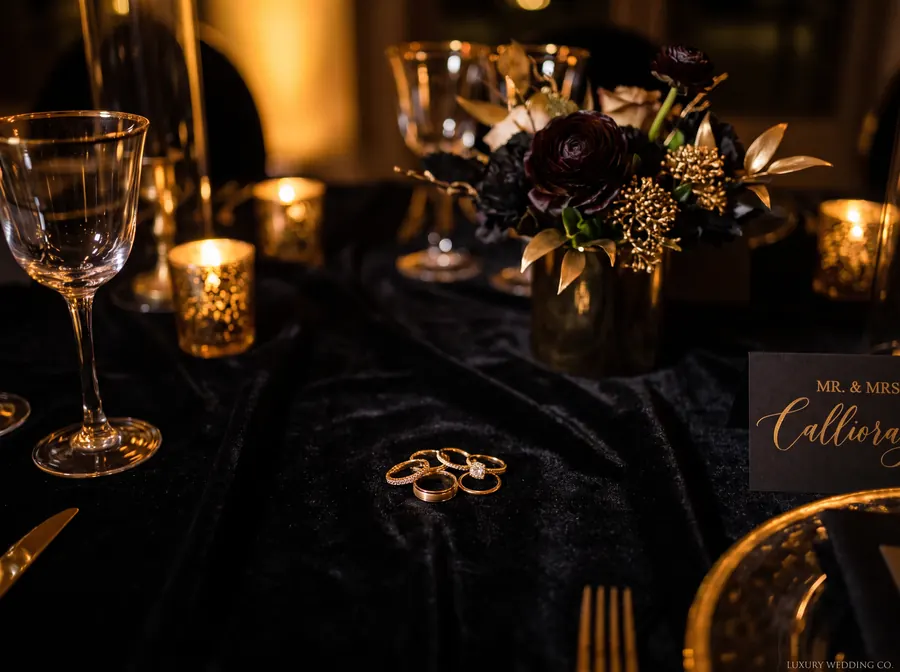 Close-up of wedding rings resting on dark velvet beside a floral centerpiece with gold accents